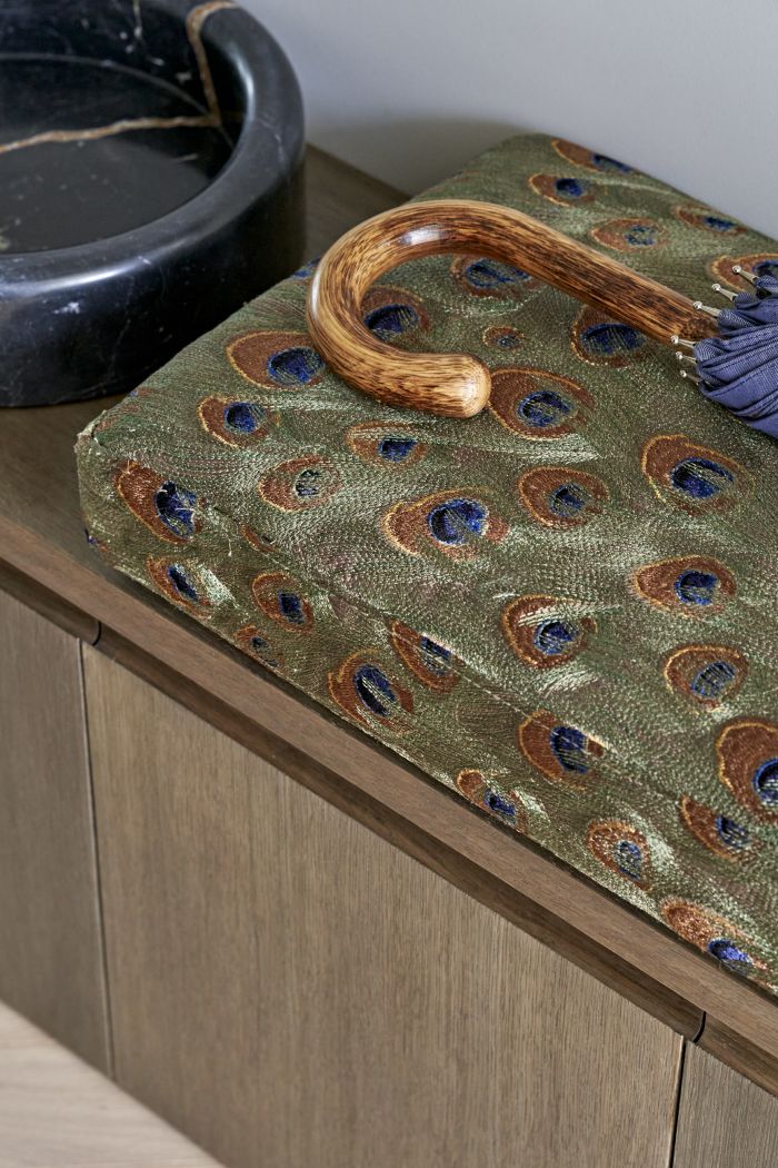 An up-close image of a luxurious area. A dark wood bench with drawers exudes functionality. Adorned with a plush cushion made out of a stunning peacock feather pattern. Alongside the cushion rests a wooden-handled umbrella. Adding a touch of sophistication, a black marble pet bowl sits nearby.