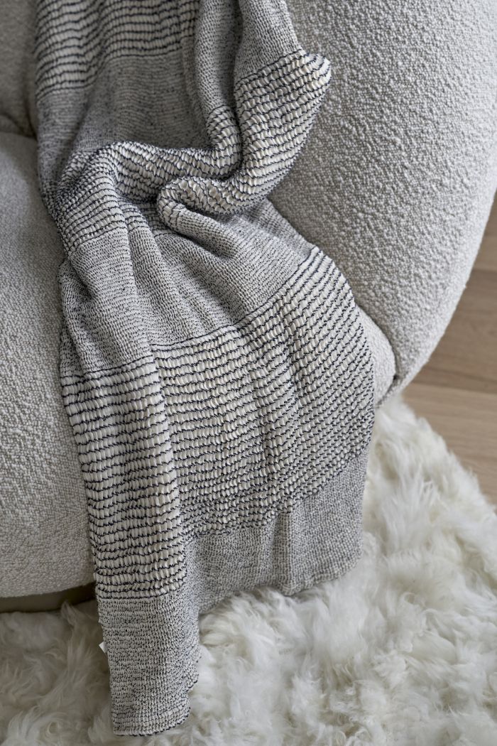 This luxurious living room captures a cozy and elegant atmosphere. A zoomed-in picture reveals a white Pacha Armchair adorned with boucle fabric, placed on a plush white fluffy rug. The light wood flooring adds warmth and sophistication to the space. A stylish throw in shades of grey and white enhances the comfort of the armchair.