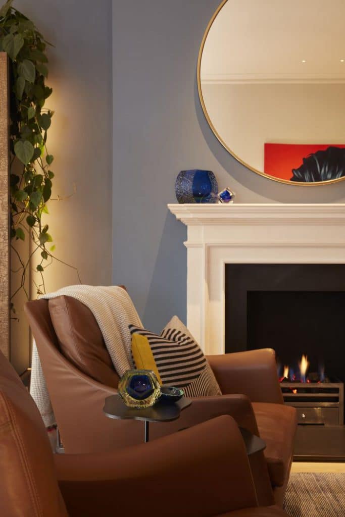 This luxury living room exudes a classic and refined ambiance. Two brown leather armchairs with striped grey and blue cushions, adorned with a white throw, create a cosy seating area. Light wood flooring complements the space, enhanced by a striped rug that adds texture and visual interest. The light grey painted walls provide a neutral backdrop, while a hanging green plant in one corner brings a touch of nature indoors. A Chesney limestone fireplace with a white mantel becomes a focal point, adorned with carefully placed ornaments. A large circular mirror above the fireplace adds depth and reflects the room's elegance.