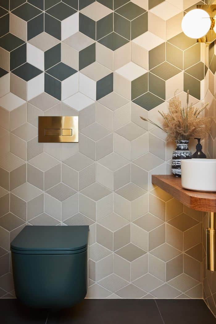 This luxurious bathroom features a avocado dark green toilet with a gold flush plate. The floor is adorned with sleek black tiles, providing a striking contrast. The walls are adorned with geometric tiles in a tasteful combination of beige and blue, adding a touch of sophistication. A wooden countertop holds a white bowl sink, accompanied by vases filled with dried flowers, bringing a natural and elegant element to the space.