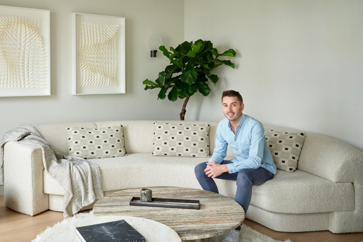 This luxurious living room features a corner space adorned with a curved white, textured sofa. Three matching white cushions and a white throw add comfort and elegance. Sitting on the sofa is renowned interior designer Brian Woulfe, responsible for the captivating design of this room. A fluffy curved rug lies in front of the sofa, hosting two marble tables in different natural tones. Adjacent to the sofa, two black side tables display books and framed photos. Adorning the wall are two white textured art pieces, while behind the sofa stands a tall, vibrant green plant, adding a touch of nature to the space.