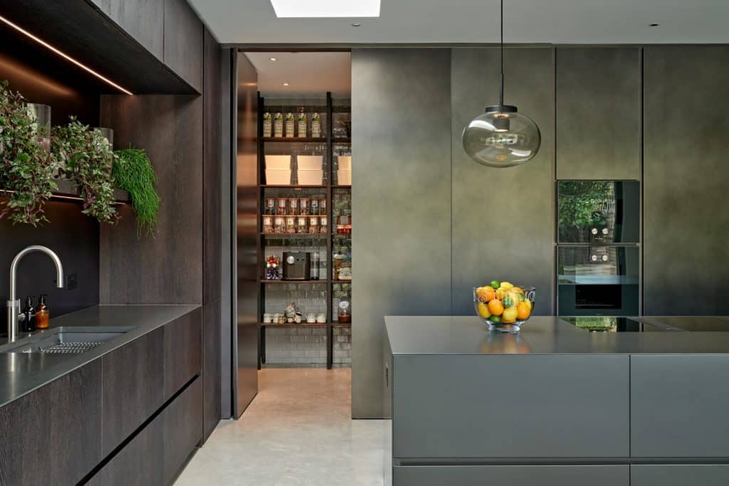 This luxury kitchen features light grey stone-look flooring, providing a sleek and sophisticated foundation. Dark wood kitchen cabinets and joinery, paired with a slate black countertop and sink, create a striking contrast. Above the sink, shelves adorned with lush plants add a touch of freshness to the space. On the opposite wall, slate black cabinets and a well-organised pantry door enhance functionality and organisation. A central slate black island houses hobs and is topped with a large glass bowl filled with vibrant oranges, lemons, and limes. Above the island, a grand glass bulb light serves as a captivating focal point.