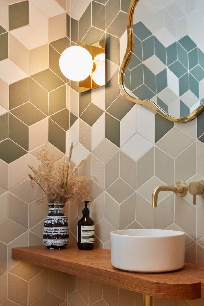 This luxury bathroom showcases a stylish and contemporary design. The walls are adorned with geometric beige and green tiles, adding a touch of sophistication. A Tom Falkner Lily mirror hangs on the wall, beautifully complemented by a gold and white globe wall light. The wooden countertop with a white bowl sink and gold tap exudes elegance, with a vase of dried flowers and a soap dispenser adding a charming touch. The combination of geometric patterns, metallic accents, and natural elements creates a luxurious and inviting atmosphere in this bathroom