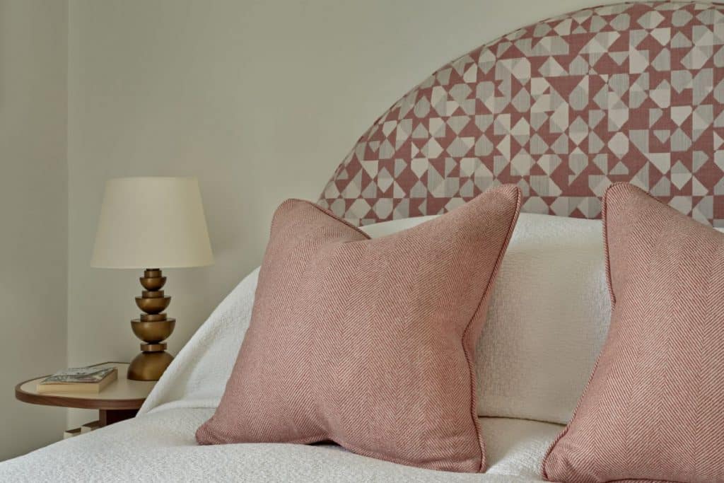 A luxury bedroom close-up image. The focal point is a magnificent bed with a curved pink and white geometric headboard, crafted from Holland and Sherry fabrics. Adorned with crisp white bedsheets and two small pink scatter cushions. Beside the bed, a circular wooden side table adds a touch of natural beauty, adorned with a book and an abstract lamp with a gold base.