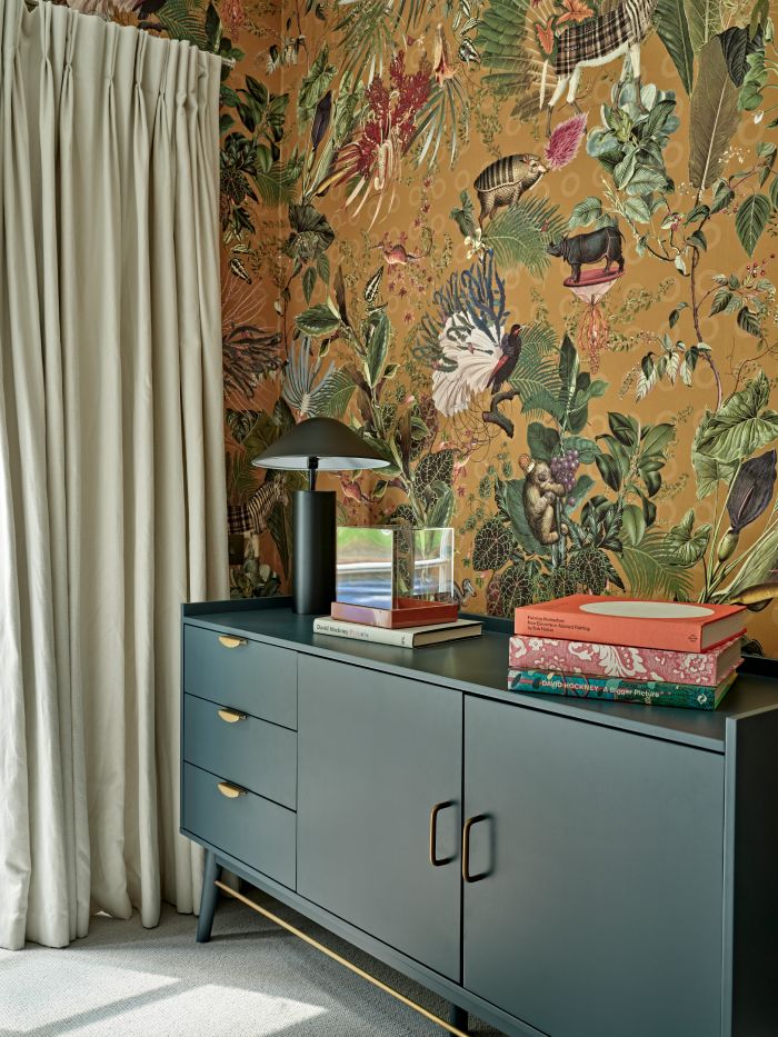 Close up image of an eye catching corner within the bedroom, where luxury meets creativity. The cream carpet and tall curtains provide a soft and elegant canvas. The wall captures attention with its unique wallpaper from 'Arte International,' adorned with a captivating display of animals, plants, and flowers. The centrepiece, a dark green cabinet with intricate gold details. Its pull-out drawers and swinging doors offer both practicality and style. Adorning the cabinet are a selection of colourful books, adding a touch of charm, sitting beside a dark green lamp.