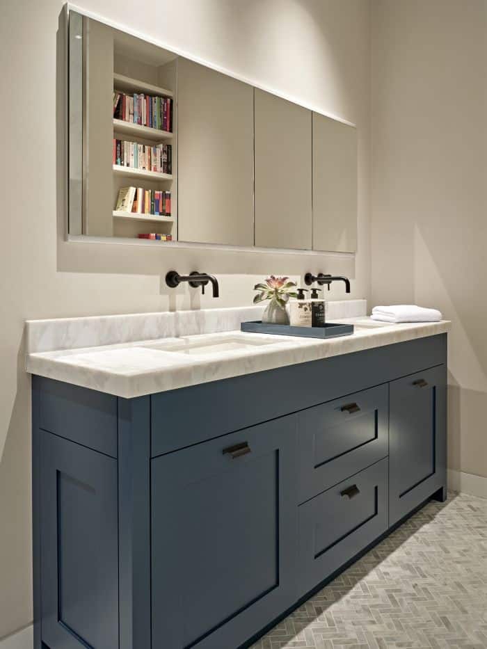 A luxurious bathroom that exudes elegance and functionality. The centrepiece of the space is a his and hers sink, with a pristine white marble countertop resting upon a beautifully crafted blue painted base. The sink is accompanied by pull-out drawers with lots of storage, ensuring a clutter-free environment. Black taps and stylish bathroom accessories add a touch of sophistication. Above the sinks, four panelled mirrors create a captivating display, reflecting the opposing wall adorned with shelves filled with a vibrant collection of colourful books.