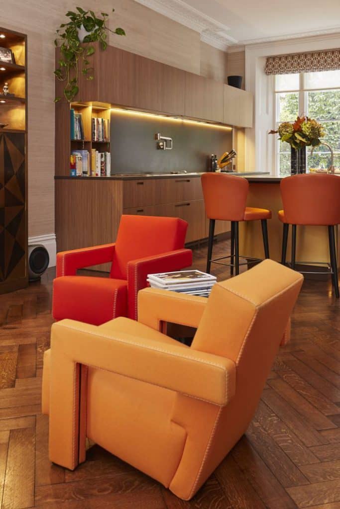 A luxurious open plan living room and kitchen.The herringbone wooden flooring sets the stage for a pair of Cassina Utrecht armchairs in bold red and orange , creating a vibrant focal point. As you glance beyond, you'll find a beautifully designed kitchen with light wood joinery and sleek grey stone countertops. Built-in cookbook shelves and a convenient pot filler add practicality to the space. At the heart of it all, a circular island with a yellow base and grey stone top, adorned with a vase of fresh flowers. Surrounding the island, orange chairs with black legs provide comfortable seating. Finally, a large window at the back floods the room in natural light.