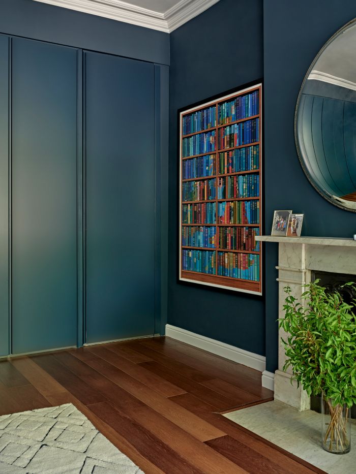 An up close image of a luxurious corner of a bedroom. The dark wood floor is adorned with a plush white rug, providing a soft and inviting touch. The teal-painted walls add a vibrant pop of colour, cleverly concealing the wardrobe doors. On one wall, a captivating painting of a bookshelf filled with brightly coloured books, enclosed in a sleek black frame, adds a touch of sophistication. Adjacent to the painting, a white marble fireplace proudly displays an assortment of picture frames, while a glass vase with lush green foliage brings a refreshing natural element to the space. Above the fireplace, a large circular mirror with a subtly curved surface creates an intriguing slight visual distortion in its reflection.