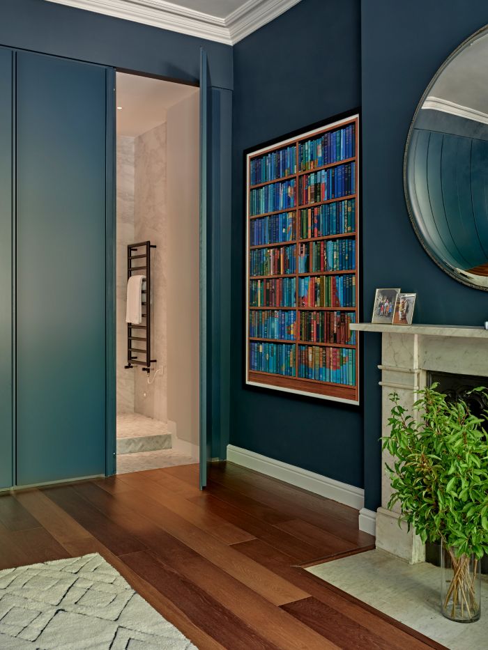 An up close image of a luxurious corner of a bedroom. The dark wood floor is adorned with a plush white rug, providing a soft and inviting touch. The teal-painted walls add a vibrant pop of colour, cleverly concealing the bathroom doors, one of the doors stands open to show a hidden luxury bathroom beyond. On one wall, a captivating painting of a bookshelf filled with brightly coloured books, enclosed in a sleek black frame, adds a touch of sophistication. Adjacent to the painting, a white marble fireplace proudly displays an assortment of picture frames, while a glass vase with lush green foliage brings a refreshing natural element to the space. Above the fireplace, a large circular mirror with a subtly curved surface creates an intriguing slight visual distortion in its reflection.