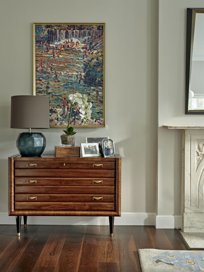 A taste of a luxurious corner of this living room, where elegance meets attention to detail. The dark wood floor sets the stage for the space, while the light green walls create a soothing canvas. A magnificent dark wood chest of drawers with gold accents takes centre stage, adorned with a display of framed photos, a delicate white orchid, and a blue-based lamp. Above the chest, a stunning colourful painting in a gold frame becomes a focal point. To the right, a glimpse of a colourful rug and a white marble fireplace.