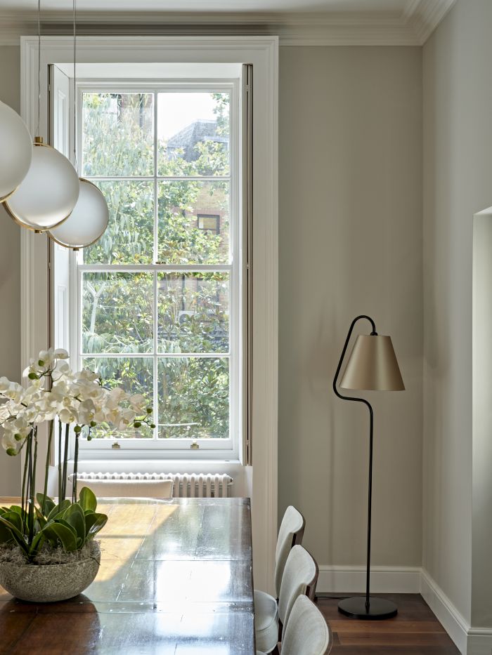 A luxurious dining room. The dark wood floor sets the stage for a beautifully varnished dark wood table which takes centre stage, adorned with delicate white orchids. Surrounding the table, elegant white chairs offer comfort and sophistication. Above, white and gold globe lights used to illuminate the space. In the background, a large window welcomes natural light, complemented by a modern floor lamp made from black metal with a golden lampshade.