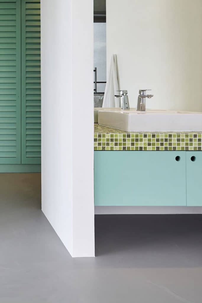 Teal green wardrobes and vanity storage in a walk in wardobe and bathroom with resin flooring.