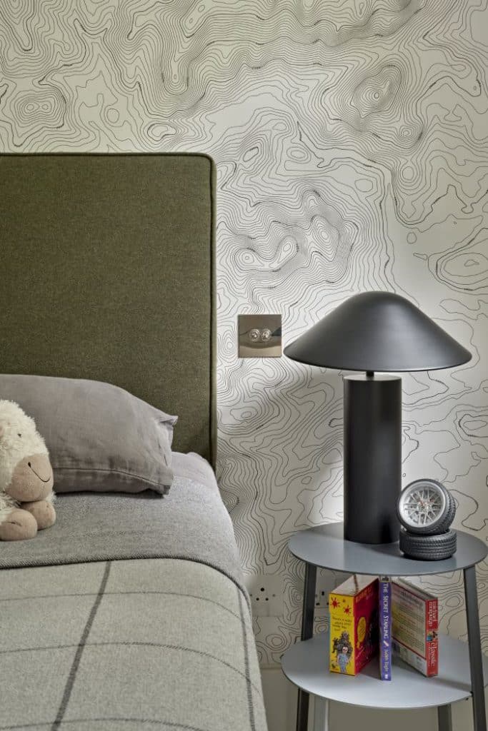 A luxurious children's bedroom, captured in a close up image. The walls are draped with a captivating black and white geographical map wallpaper, adorned with a silver light switch. A deep green headboard serves as the focal point. The bed is dressed in cosy grey sheets, accompanied by a cuddly stuffed toy resting on top. A sleek grey bedside table showcases a black bedside lamp with a whimsical mushroom-shaped lampshade. Books and accessories complete the table.