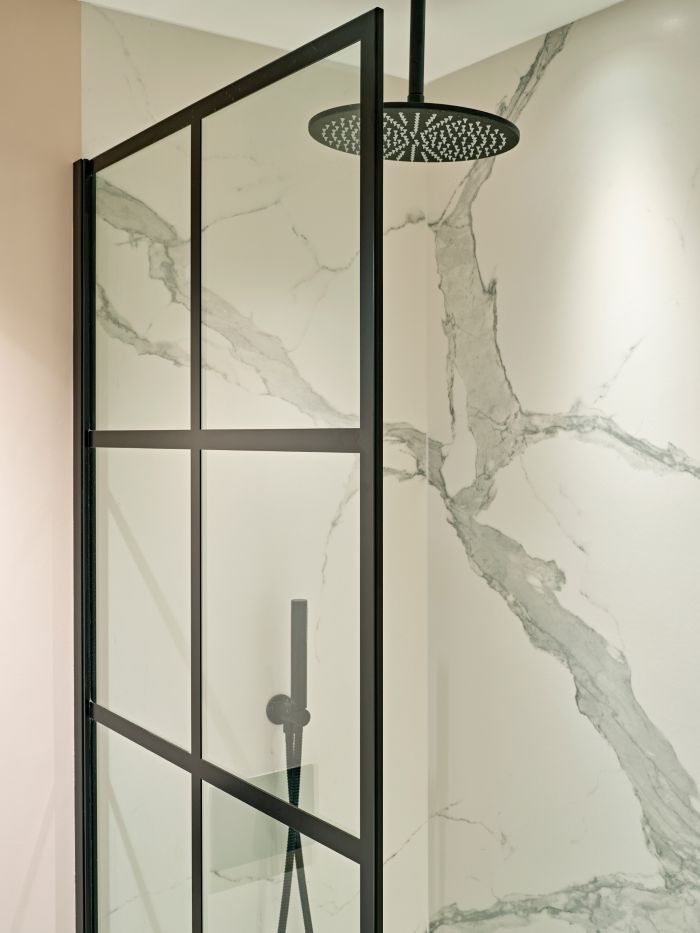 A close up image of a shower within a luxury bathroom. The shower has beautiful white marble tiles on the walls, with grey veins running through them, complemented by a black shower head and hardware. The glass screen is framed in black and split up into an elegant grid style giving the space a sophisticated touch.