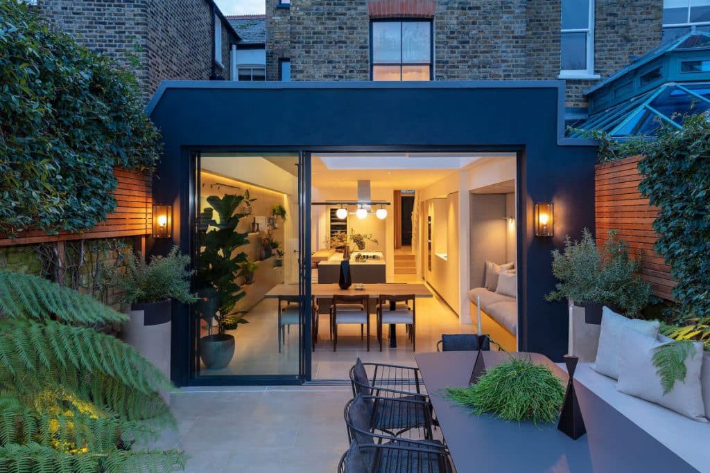 View of a dining room and kitchen from the garden. In the garden as well a dining area is located. Warm toned soft lightning sets the relaxed atmosphere of the space.