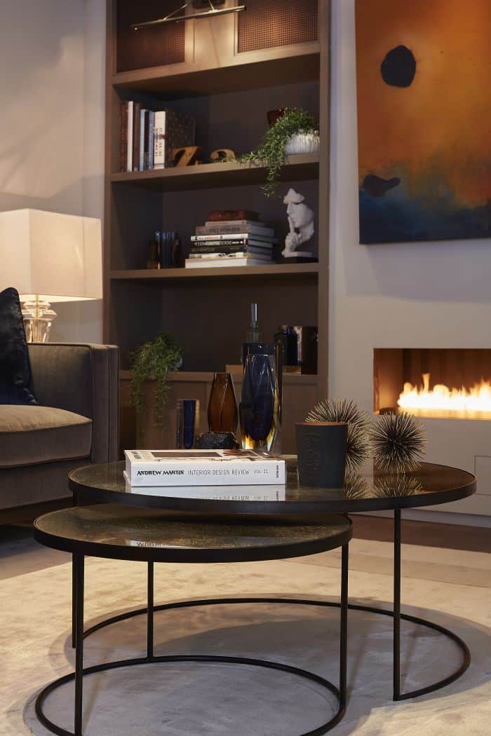 A set of nesting circular coffee tables on a rug in a living room with a lit fireplace. 