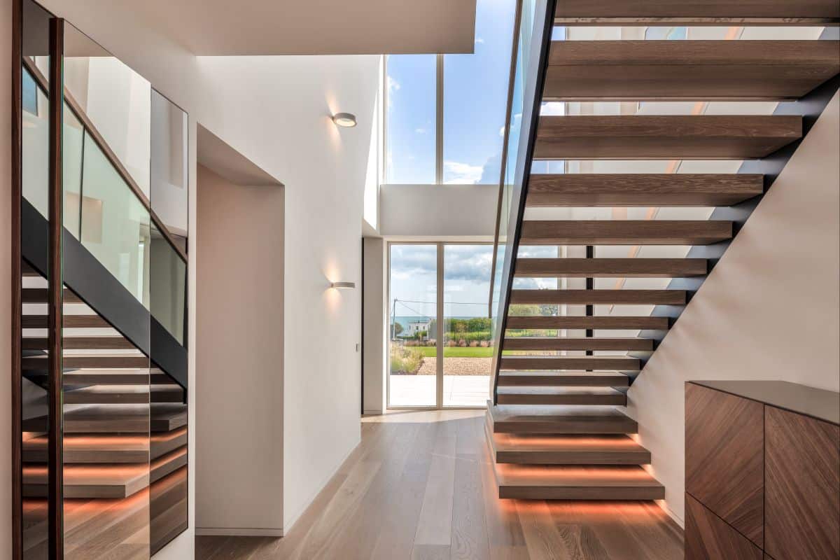 A luxury high-end hallway. The pristine white painted walls create an atmosphere of elegance, complemented by the light wood flooring. The hallway has a modern staircase, its sleek design makes a focal point for the space. Natural light streams through the large glass windows by the front door. Opposite the staircase, three grand mirrors reflect the staircase. Circular ringed lights adorning the walls.