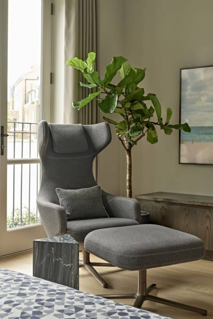 This luxurious media room/study exudes an elegant and serene atmosphere. The light wood flooring sets a warm foundation, complemented by large windows adorned with cream curtains that allow natural light to fill the space. The centrepiece is the Grand Repos armchair and ottoman by Vitra in a stylish grey hue, inviting relaxation and comfort. A wood block triangular side table stands alongside the chair, providing a convenient surface for books or beverages. A tall green plant adds a touch of nature, while a wooden ottoman with framed art above it completes the sophisticated ambience of this luxurious media room/study.